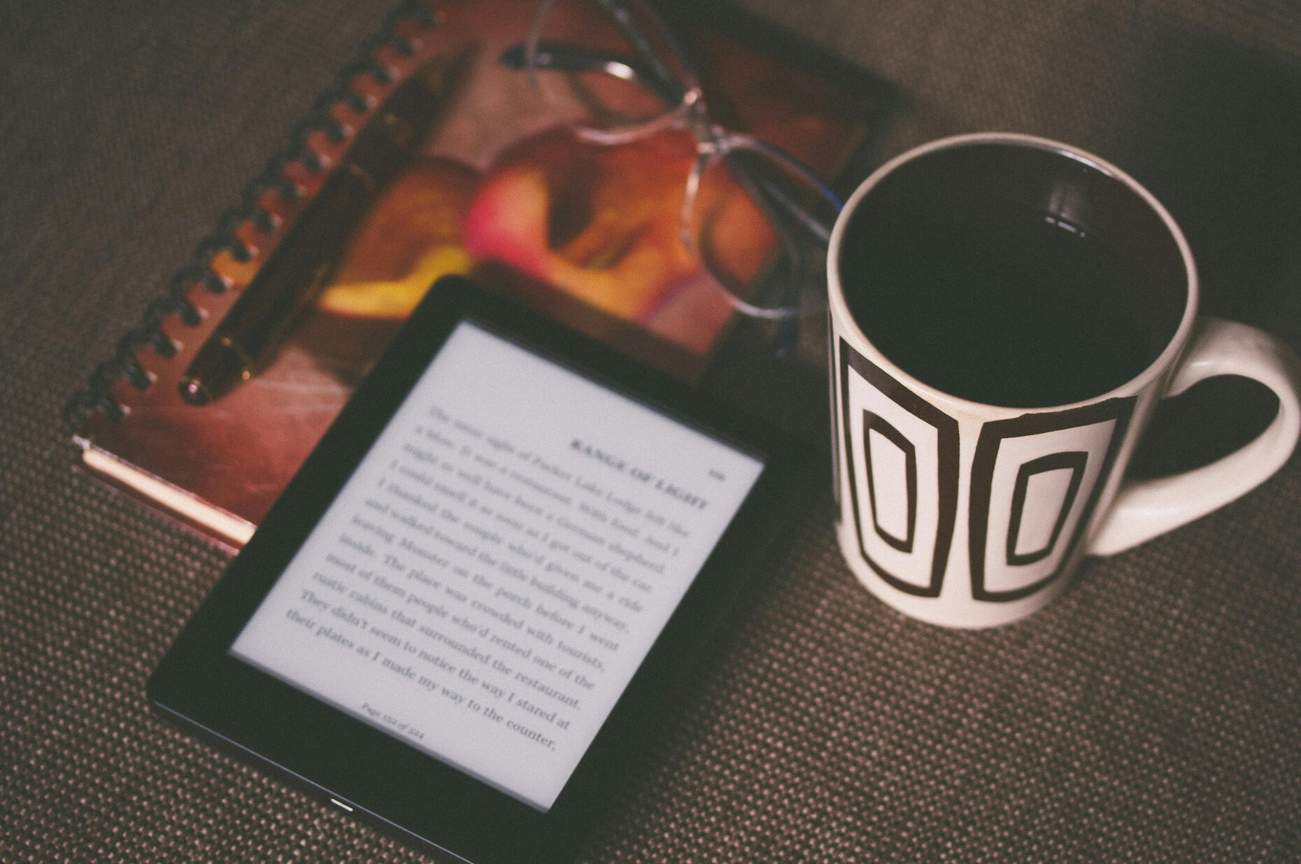 ebook reader surrounded by a coffee mug, glasses and a journal